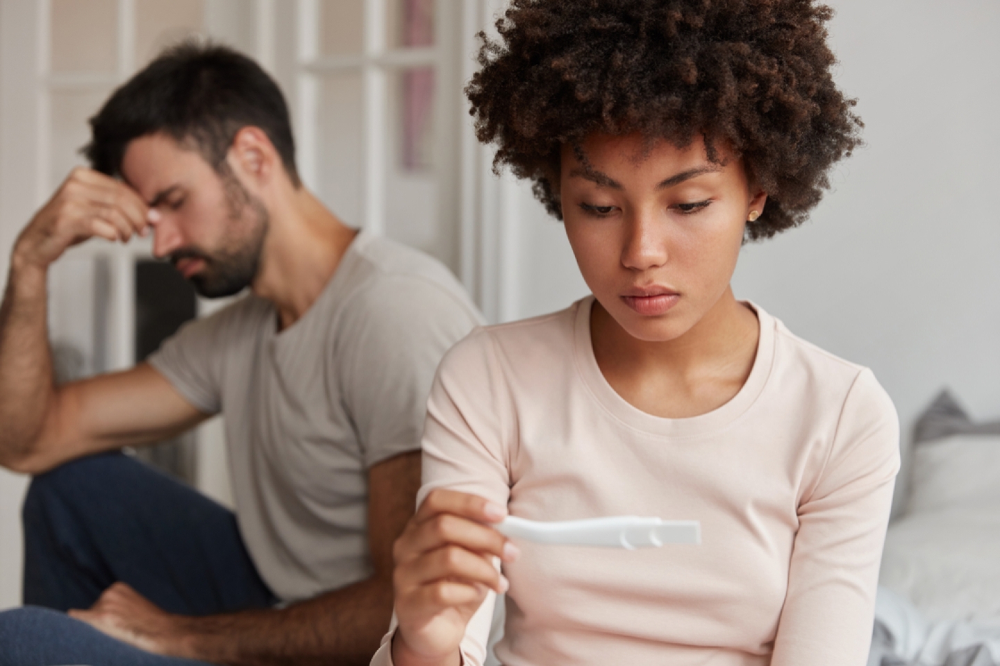 Upset dark skinned attractive woman looks at pregnancy test, suffers from infertility, frustrated husband sits near at edge of bed. Family couple have problem, cant have children for long time