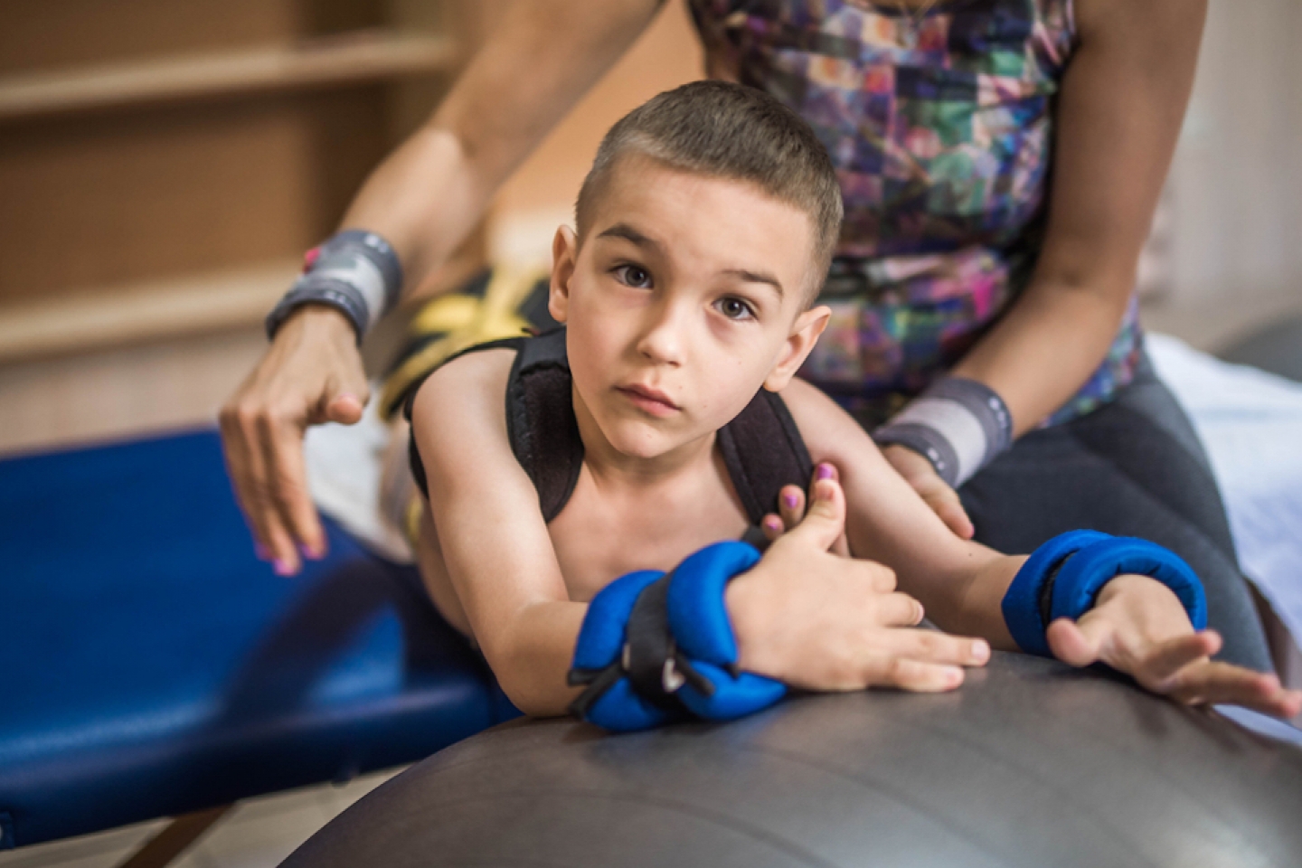 Physical therapist assisting little boy make exercises on gym ball during rehabilitation in children hospital.