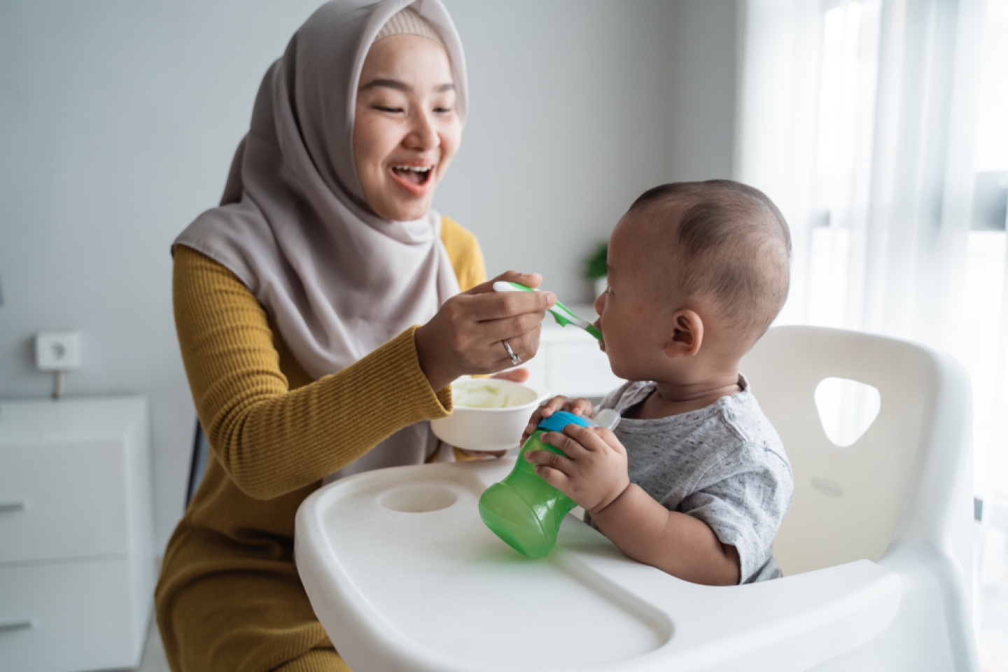 muslim asian mother feeding her baby boy while sitting on high chair at home