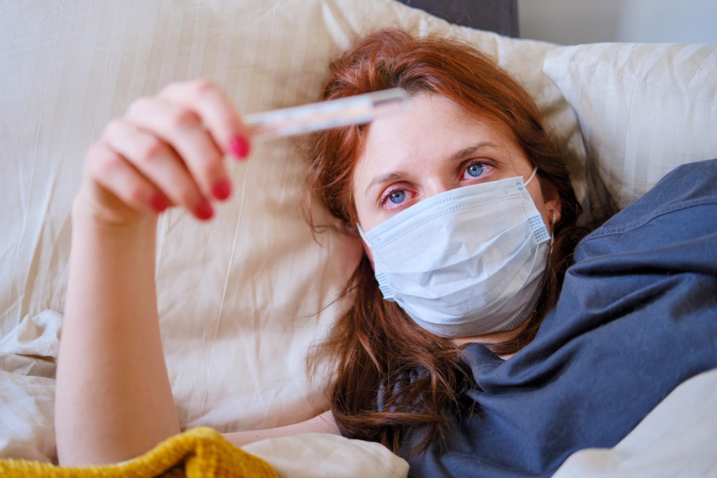 Young red-haired woman looks at a thermometer while lying in bed. Sick girl lies at home in a medical mask