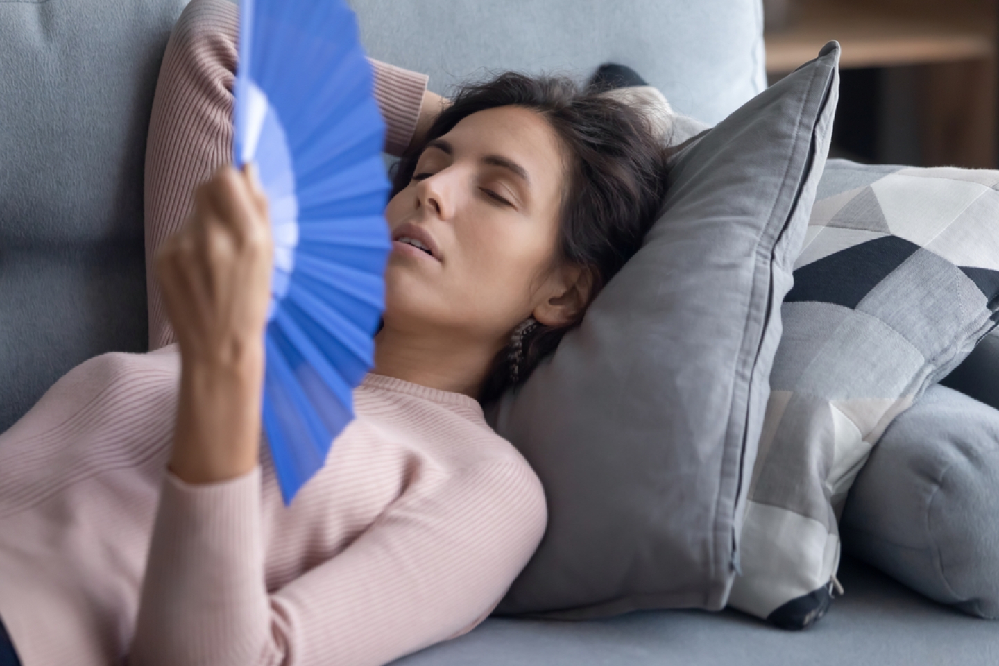 Exhausted overheated woman waving blue paper fan close up, lying on couch