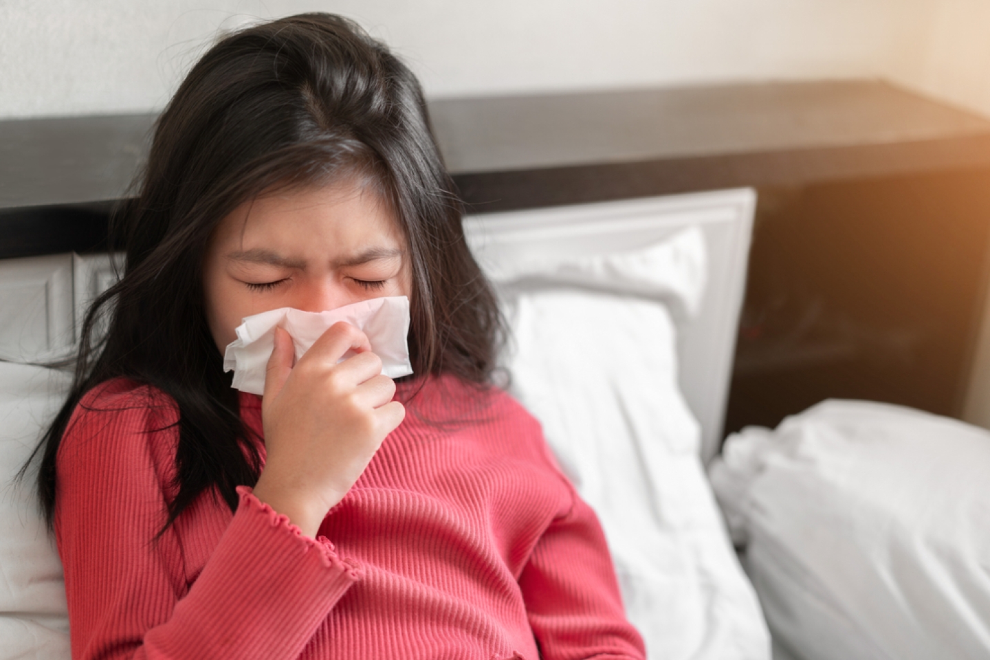 Sick girl sneezing into tissue on bed in bedroom