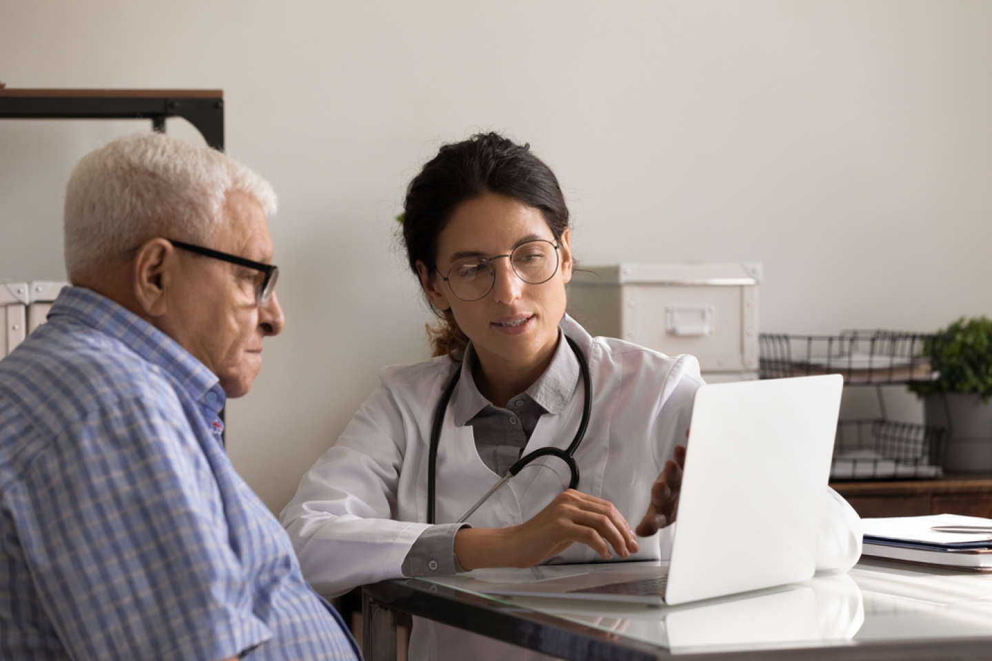 Young female doctor and senior male patient look at laptop screen discuss treatment or therapy at consultation.