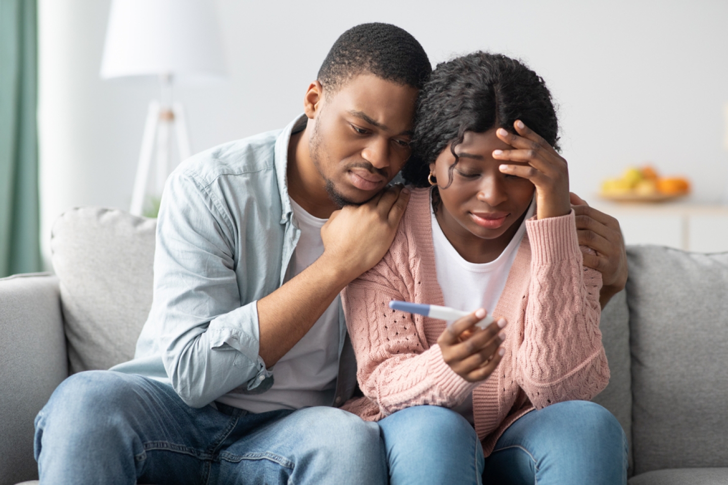 Upset black couple with negative pregnancy test sitting on couch at home, hugging and looking at test, free space.