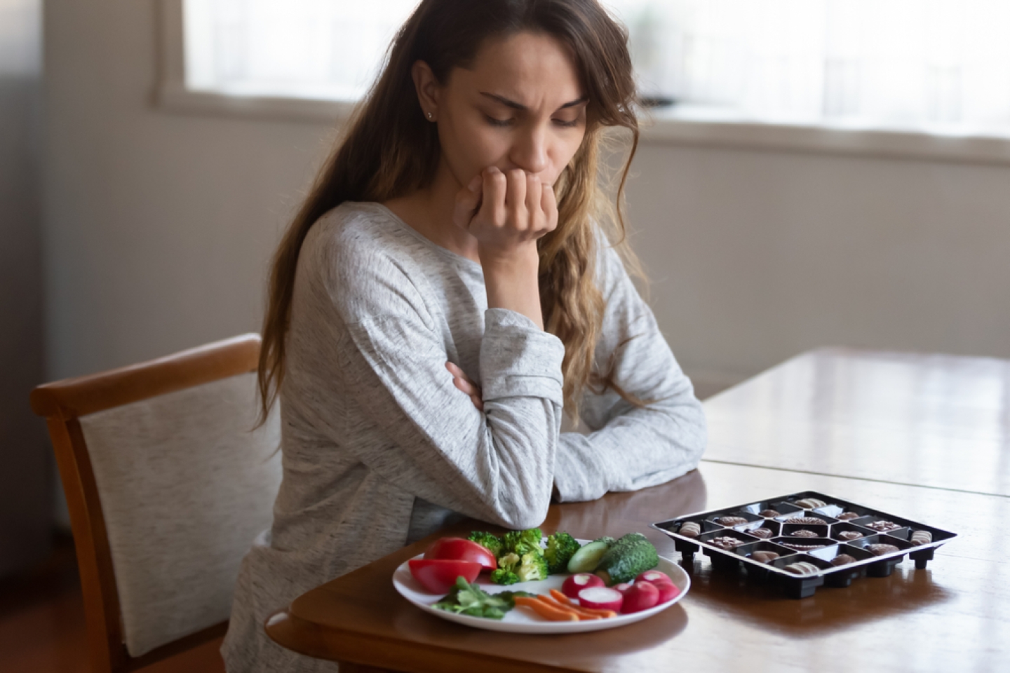 Unhappy young Latin woman look at chocolates and vegetables face temptation suffer from eating disorder.