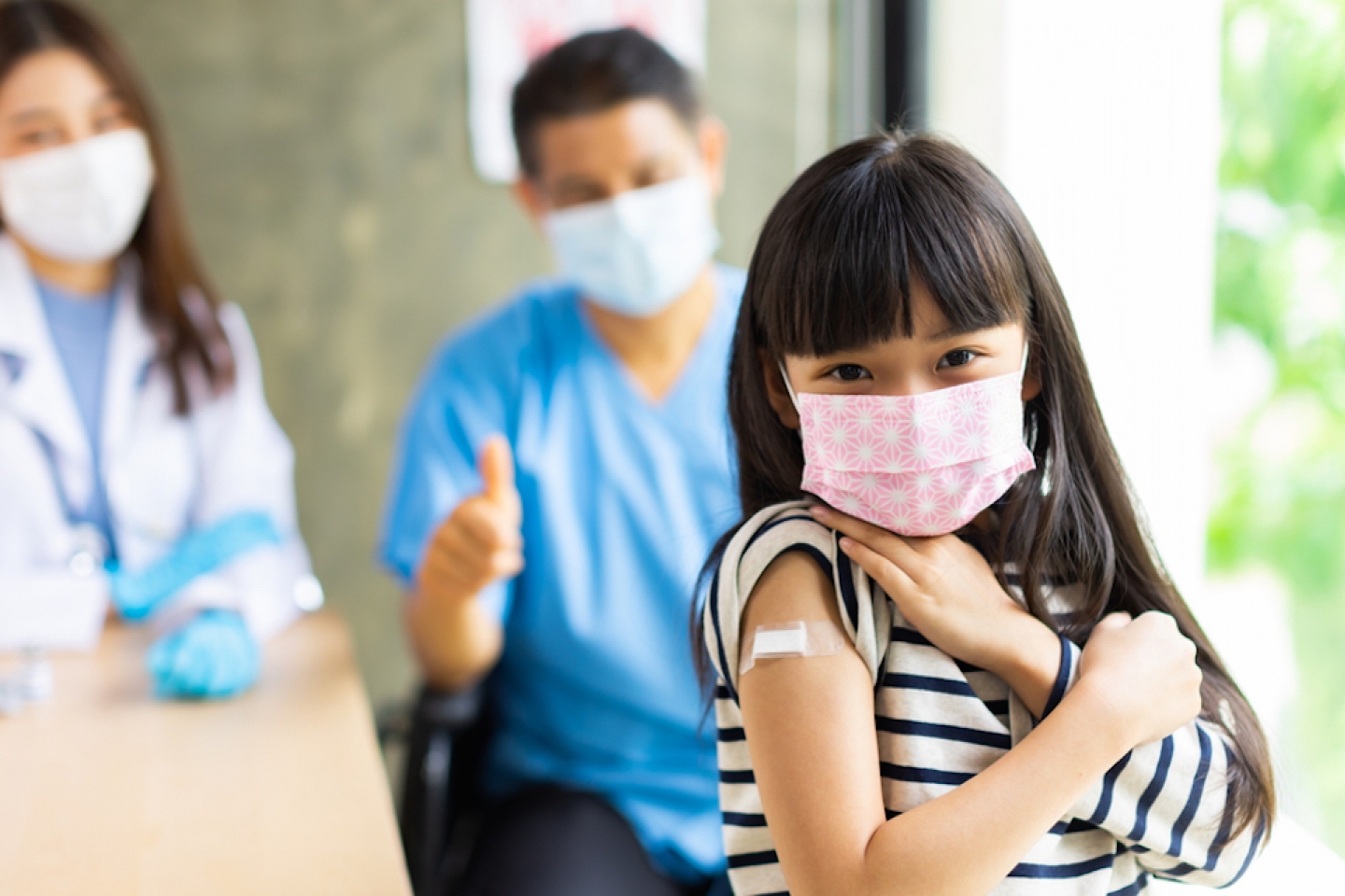 Asian doctor wearing gloves and isolation mask is making a COVID-19 vaccination in the shoulder of child patient with her mother at hospital.