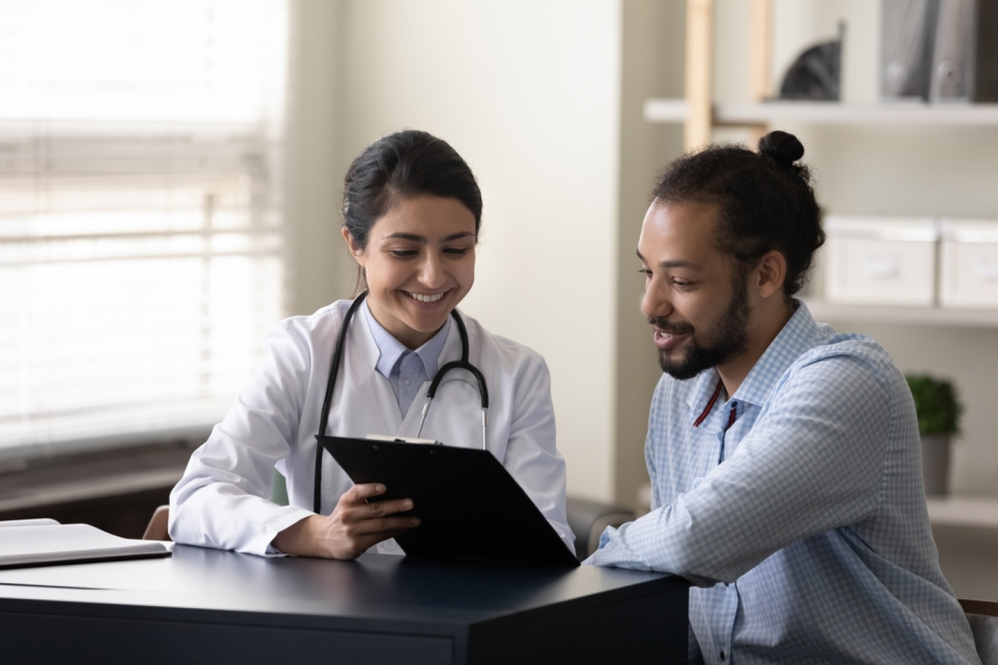 Happy young indian female gp doctor showing test result on clipboard to interested millennial african ethnicity male patient, discussing healthcare medical insurance or illness treatment at clinic.