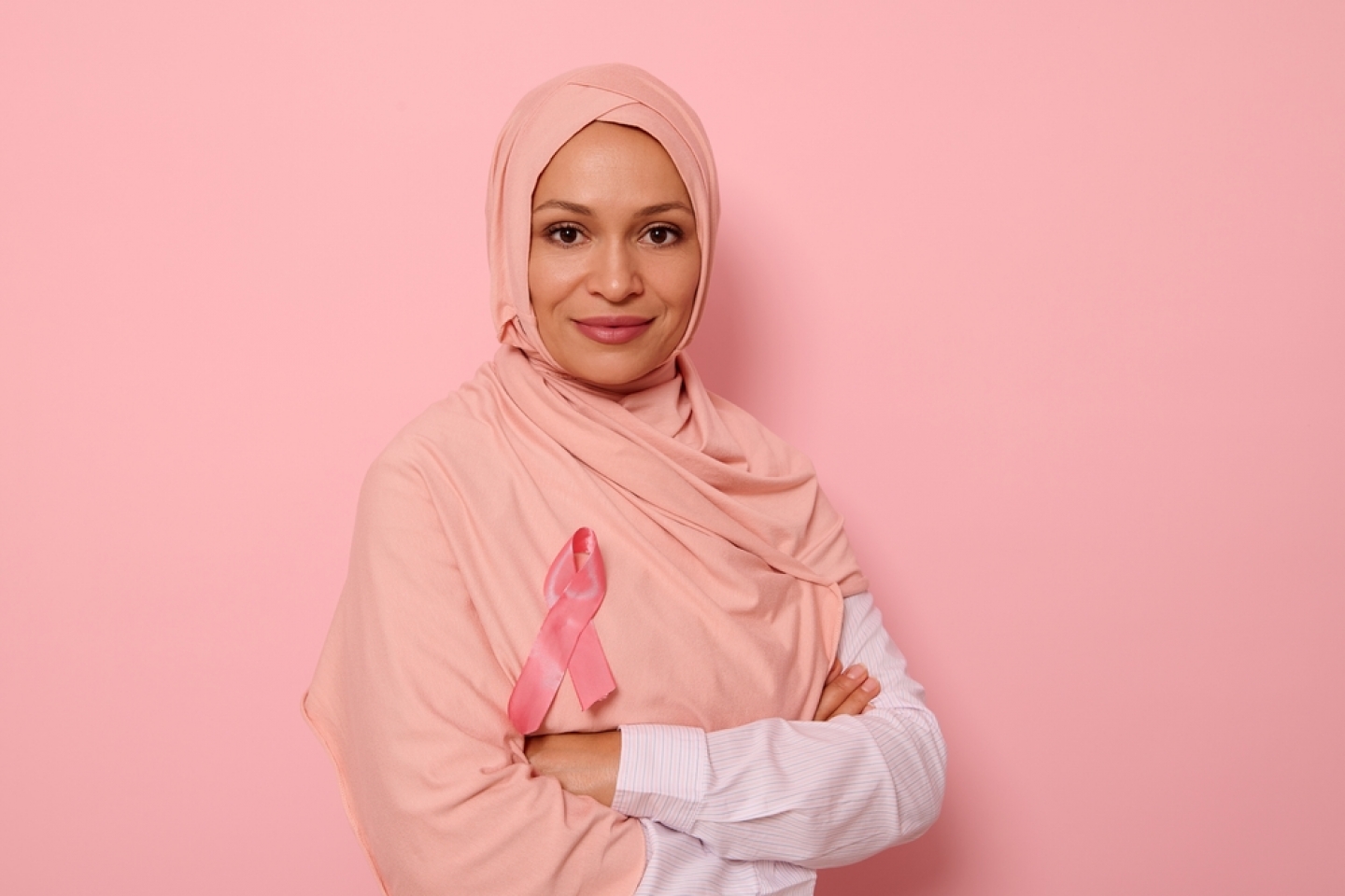 Confident portrait of a friendly Arab Muslim woman wearing a hijab and pink satin ribbon showing her support for cancer patients and survivors.