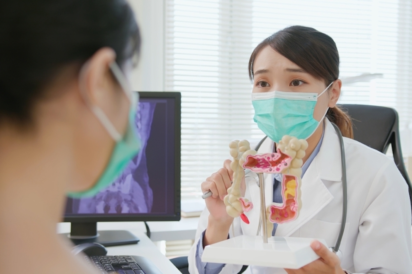 asian woman wears face mask to prevent COVID19 has colorectal cancer diagnosis in hospital - female doctor showing colonoscopy results and xray to patient on computer and explain by anatomical model