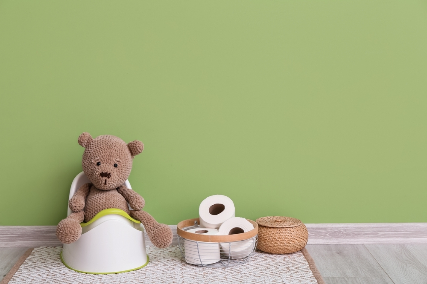 Cute toy bear sitting on potty and basket with toilet paper rolls on grey wooden floor near color wall