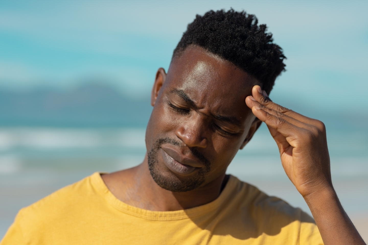 Close-up of african american young man with eyes closed touching head at beach against sky in summer. headache, unaltered, healthcare, pain, holiday, beach, lifestyle and sickness concept.