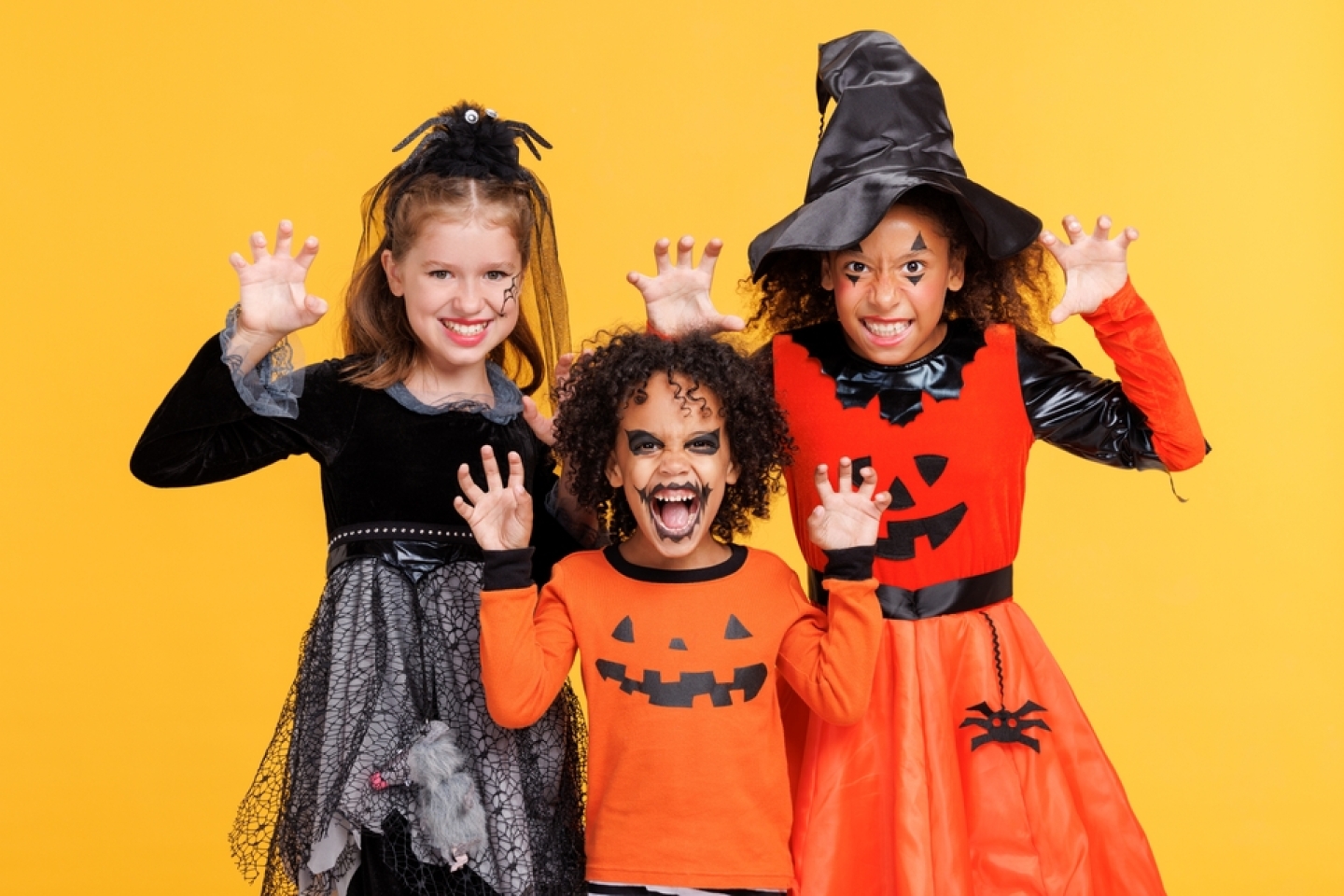 Happy Halloween! Cheerful kids in carnival costumes and makeup make a terrible gesture on bright colored yellow background