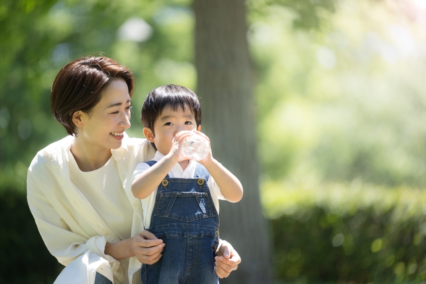 A mother gives her child a drink of water in a fresh green park. An image of hydration in the hot sun in summer. 