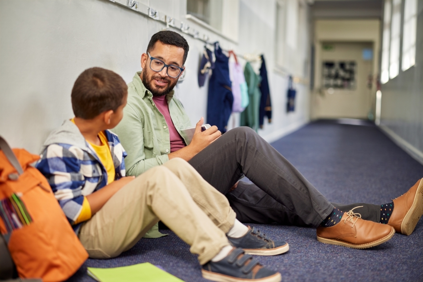 Young male psychologist speaking to depressed boy in school while sitting on the floor in school corridor.