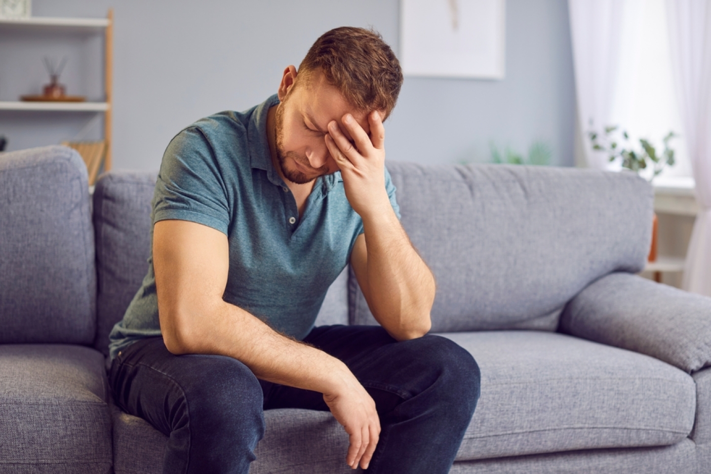 Stressed upset man sitting alone on a comfortable couch, appearing distraught, holding his head in hands, deep in thought or contemplation. Sad guy thinking about problems in relationships or at work