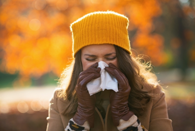 Hello autumn. elegant female in beige coat and orange hat with napkin blowing nose outdoors on the city park in autumn.
