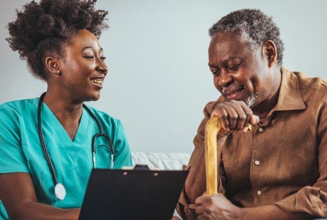 A black mid-adult nurse with a clipboard attends to an elderly black male at home, symbolizing professional healthcare and support with a comforting presence.