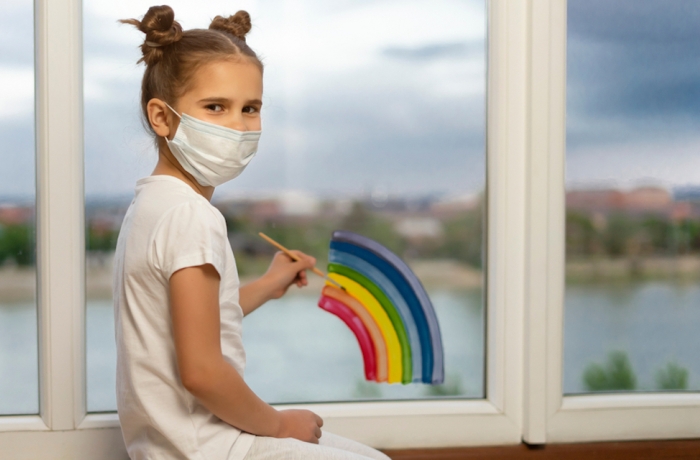 Masked schoolgirl at home draws a rainbow on the window. Girl kid create artist paints. let's all be well. Flash mob society community on self-isolation quarantine pandemic coronavirus.