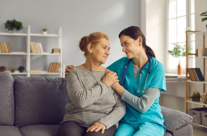 Young doctor supporting senior woman. Friendly nurse or clinician and happy older adult patient sitting on sofa, looking at each other, holding hands and smiling. Health check, trust and care concept