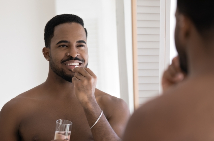 Handsome hipster African man looks in mirror caring for health takes pill hold glass of still water.