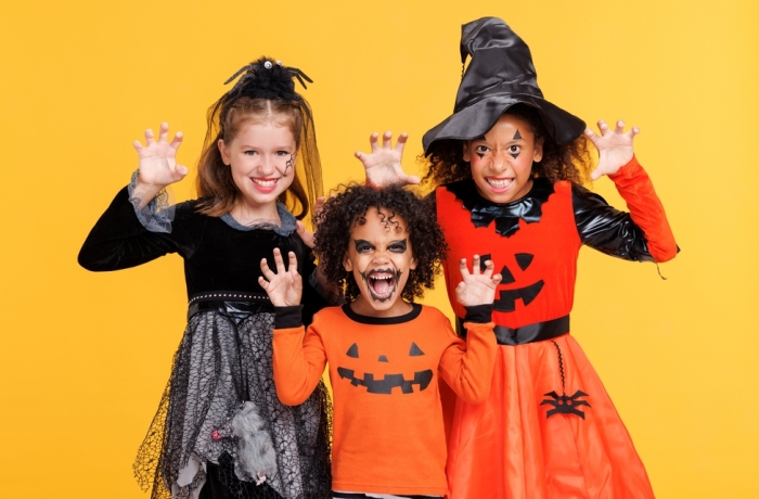 Happy Halloween! Cheerful kids in carnival costumes and makeup make a terrible gesture on bright colored yellow background