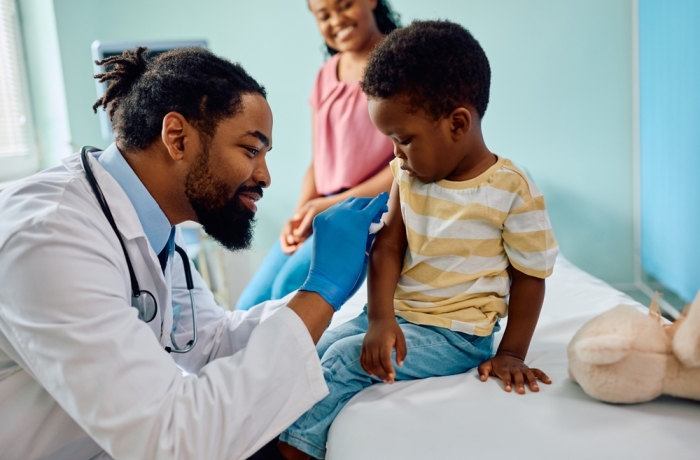 Caring African American pediatrician preparing arm of a small boy for vaccination at doctor's office.