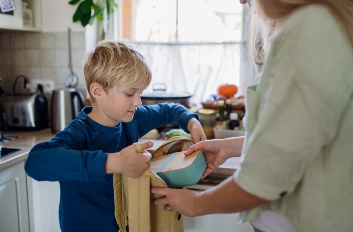 Mom packing snack for her son for school. Putting lunch box with healthy snack into his school bag for lunch.