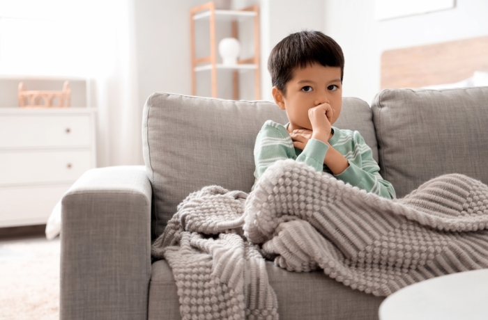 Ill little Asian boy coughing on sofa at home