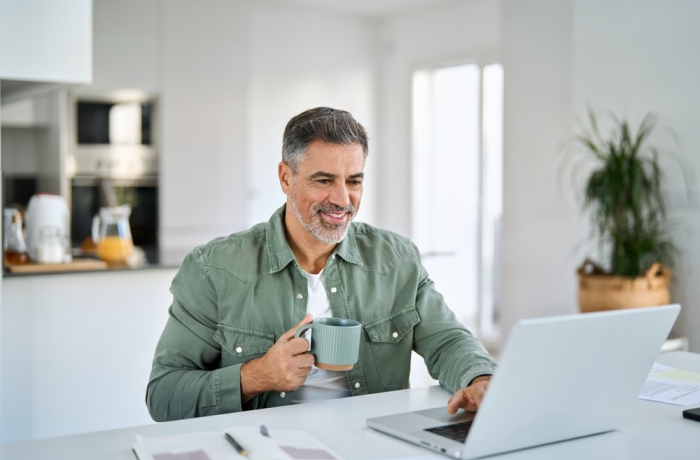 Smiling happy older mature middle aged professional man drinking coffee looking at computer sitting at kitchen table, using laptop hybrid working, elearning, browsing online relaxing at casual home.