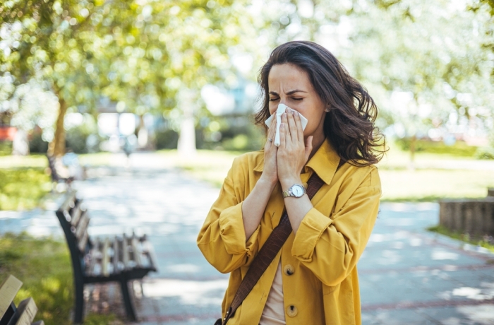 Close-up of a young woman blowing nose with tissue paper at the park. Woman with with allergy symptom blowing nose. Young pretty woman sneezing in front of blooming tree. Spring allergy concept