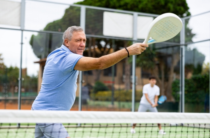 Focused aged man playing friendly paddleball match on outdoor summer court. Senior people sports concept..