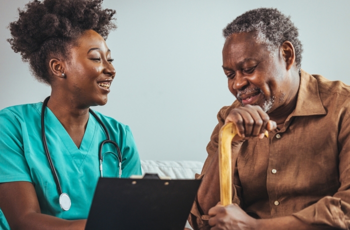A black mid-adult nurse with a clipboard attends to an elderly black male at home, symbolizing professional healthcare and support with a comforting presence.