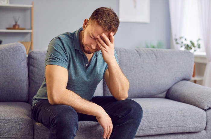 Stressed upset man sitting alone on a comfortable couch, appearing distraught, holding his head in hands, deep in thought or contemplation. Sad guy thinking about problems in relationships or at work