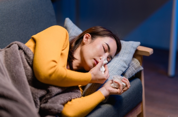 This shows an Asian woman who is sick resting on a sofa using tissues She is dealing with a cold or flu and seeks remedy and relief Healthcare is important
