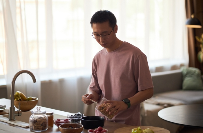 Young adult Asian man preparing breakfast in modern kitchen, pouring granola into bowl with fresh fruit on counter, standing near sink with natural light coming through window