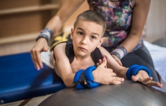Physical therapist assisting little boy make exercises on gym ball during rehabilitation in children hospital.