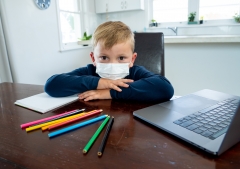 child with face mask on at desk with laptop, paper and coloring pencils