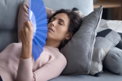 Exhausted overheated woman waving blue paper fan close up, lying on couch