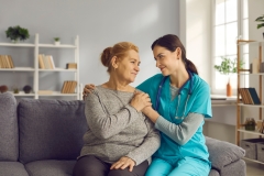 Young doctor supporting senior woman. Friendly nurse or clinician and happy older adult patient sitting on sofa, looking at each other, holding hands and smiling. Health check, trust and care concept