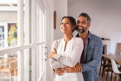 Smiling mid adult couple hugging each other and standing near window while looking outside.