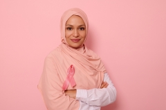 Confident portrait of a friendly Arab Muslim woman wearing a hijab and pink satin ribbon showing her support for cancer patients and survivors.