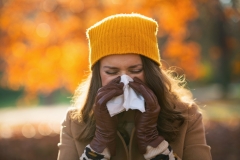 Hello autumn. elegant female in beige coat and orange hat with napkin blowing nose outdoors on the city park in autumn.