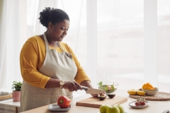 Portrait of young black woman cutting avocado while making salad in sunlit kitchen,