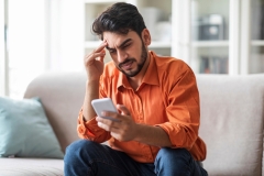 Stressed upset man in casual sitting on couch at home, holding cell phone