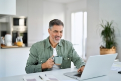 Smiling happy older mature middle aged professional man drinking coffee looking at computer sitting at kitchen table, using laptop hybrid working, elearning, browsing online relaxing at casual home.