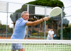 Focused aged man playing friendly paddleball match on outdoor summer court. Senior people sports concept..