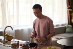 Young adult Asian man preparing breakfast in modern kitchen, pouring granola into bowl with fresh fruit on counter, standing near sink with natural light coming through window